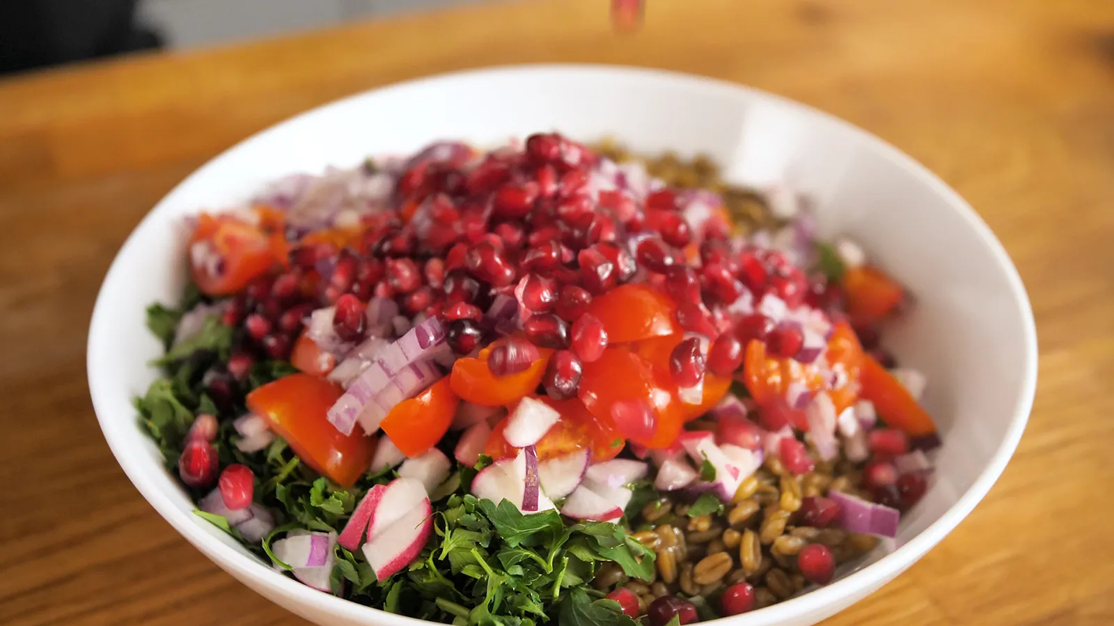 A hand sprinkles fresh pomegranate seeds over a white bowl filled with a colorful salad of wheat berries, chopped vegetables, and herbs.