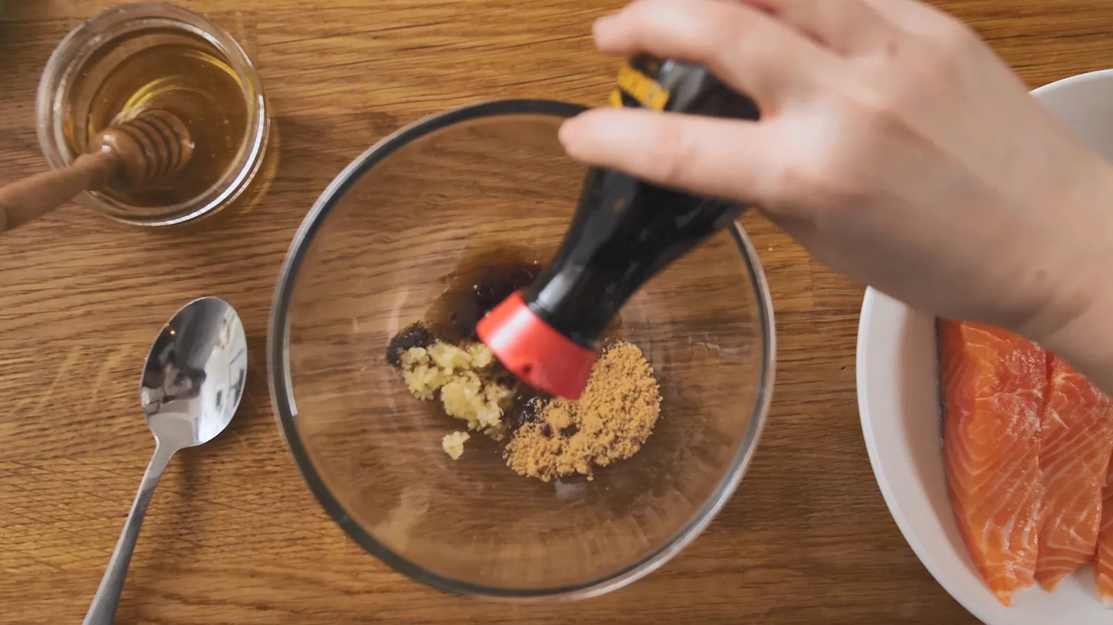A hand pours soy sauce into a glass bowl containing minced garlic and brown sugar, preparing a marinade for salmon fillets.