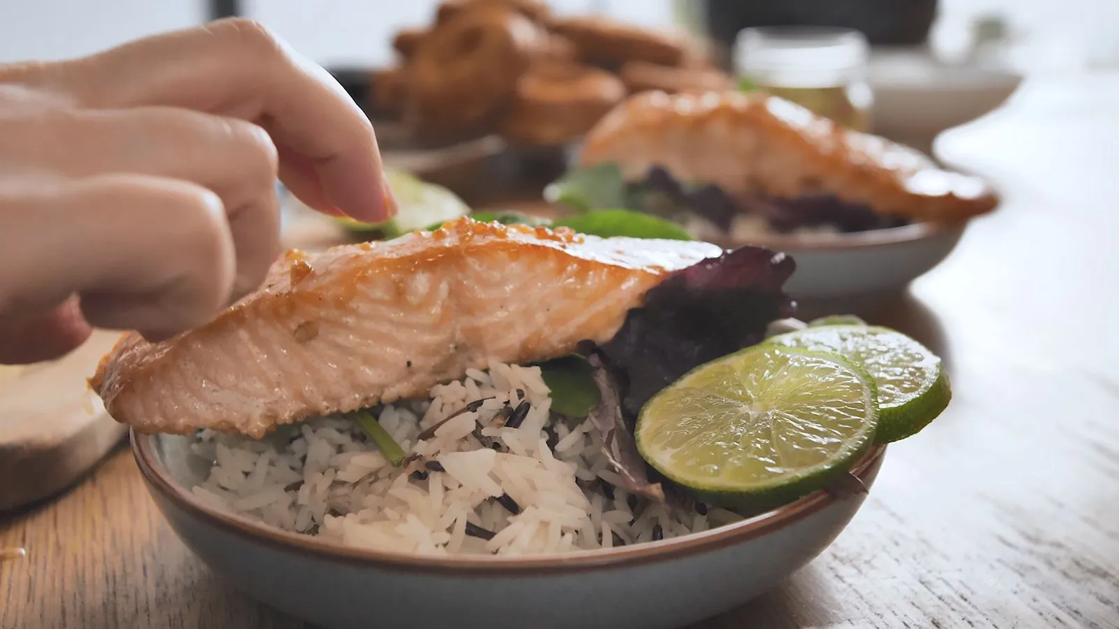 A hand places a cooked salmon fillet on a bowl of rice and mixed greens, garnished with lime slices, as part of a plated meal.