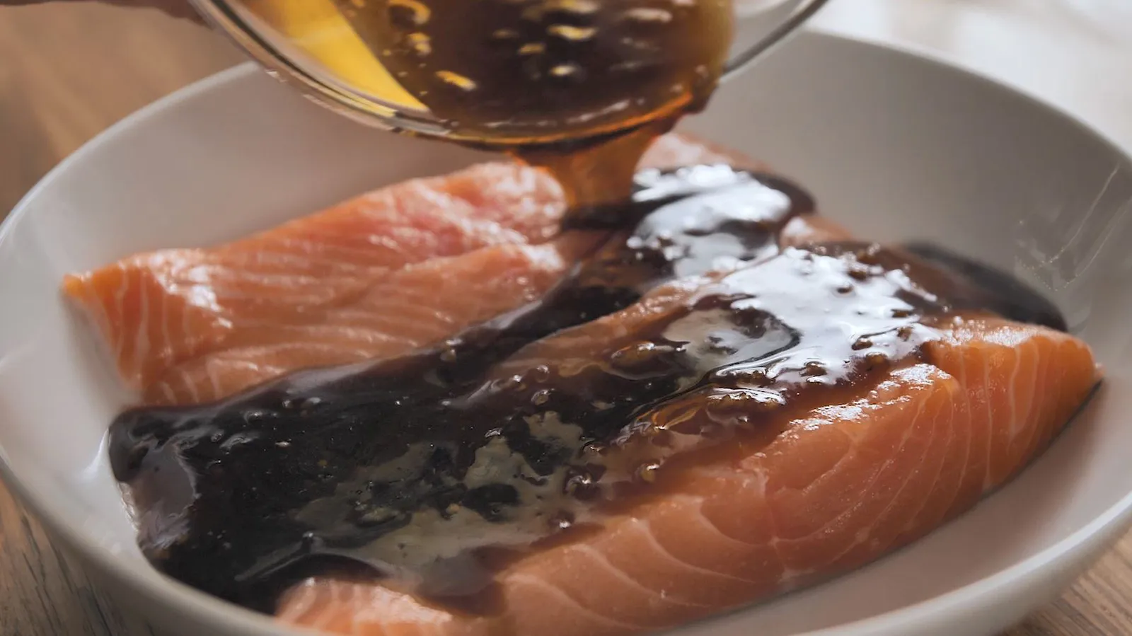 A dark marinade is poured from a glass bowl over raw salmon fillets in a white dish, preparing them for cooking.