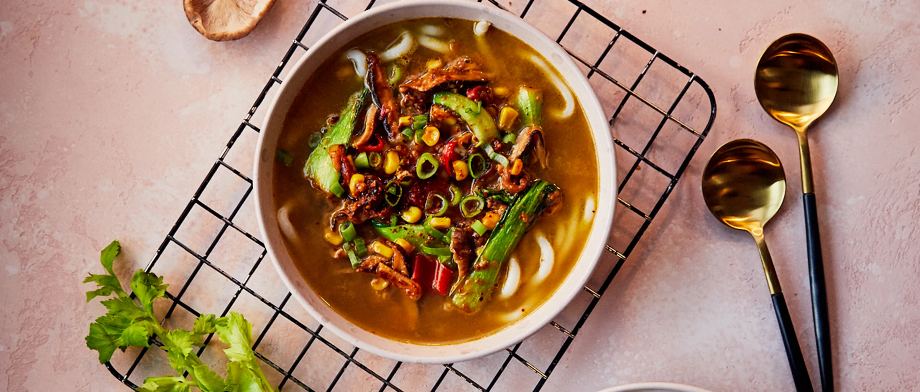Hearty vegetable soup with noodles, spices, and garnishes on a pink background.