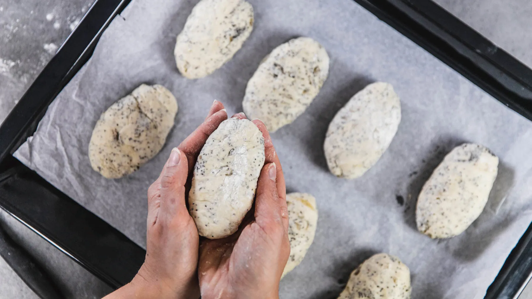 Hände formen ein längliches Brötchen aus Teig auf einem mit Backpapier belegten Backblech mit mehreren ähnlichen Teiglingen.