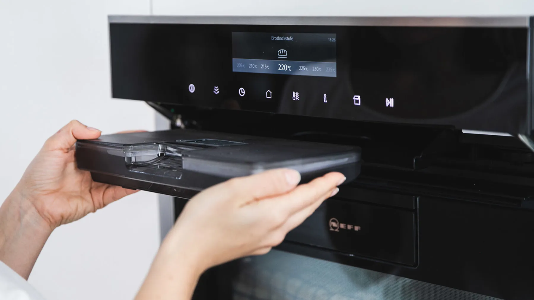 Hände schieben eine schwarze Auffangschale in einen schwarzen Backofen mit digitalem Display