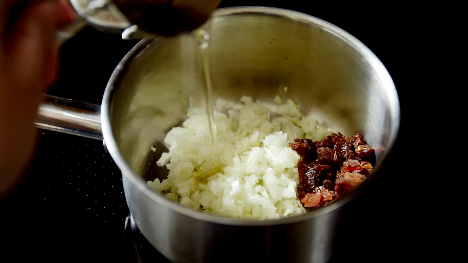 Oil is poured from a measuring spoon into a stainless steel saucepan containing diced onions and dried fruit, cooking on an induction hob.