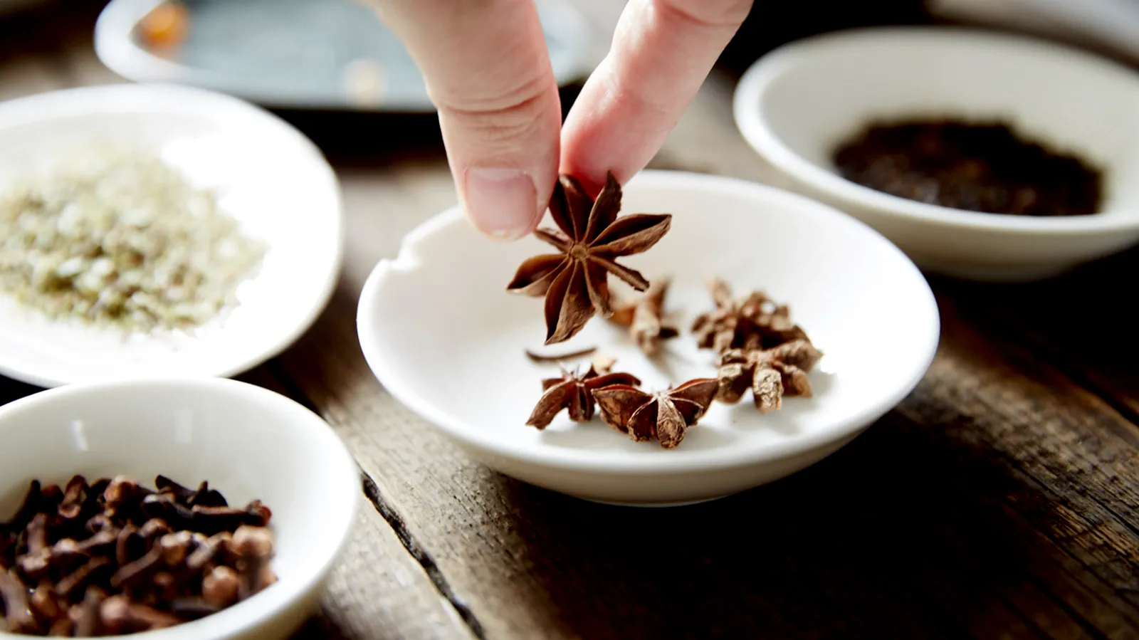 A hand picks up a star anise from a white bowl, surrounded by other spices like cloves, on a wooden table.