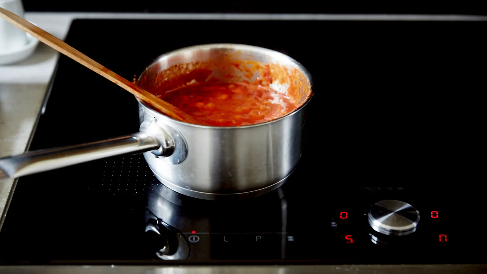 A stainless steel saucepan with red sauce is simmering on a black induction cooktop, being stirred with a wooden spoon.
