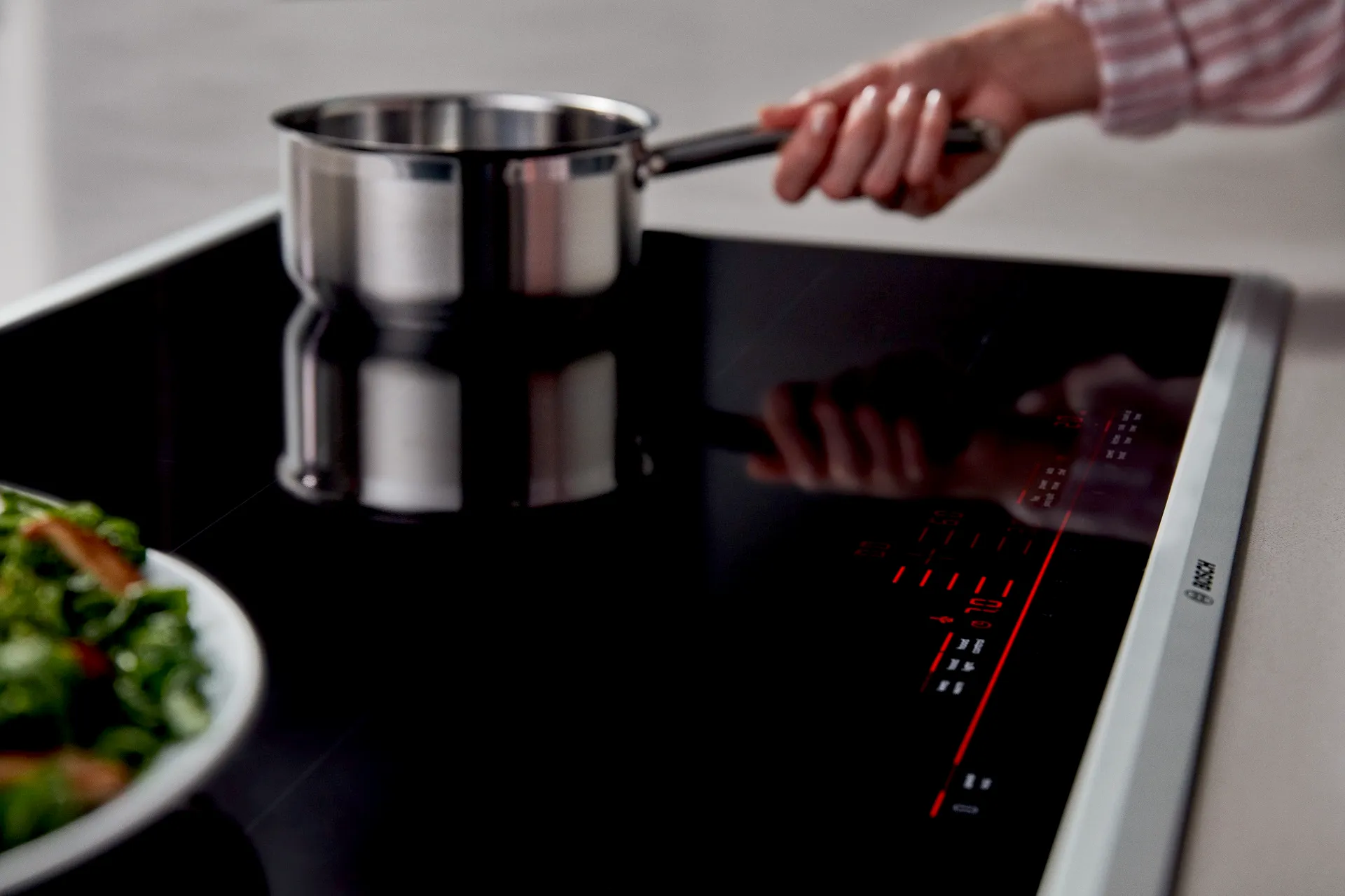 Person cooking on a sleek Bosch black induction stovetop with digital controls, holding a stainless pot.