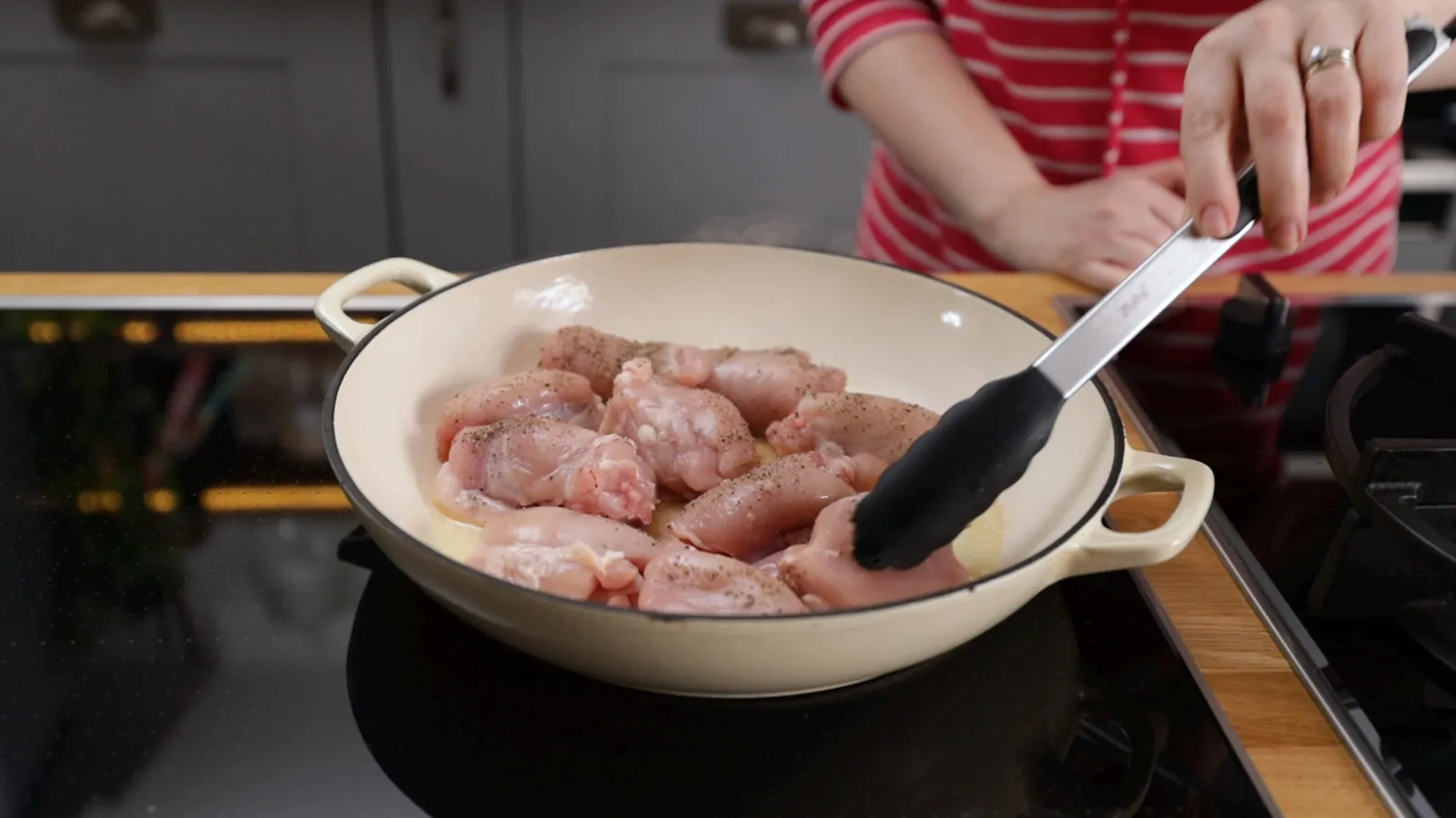 A hand uses black tongs to turn raw chicken pieces seasoned with salt and pepper in a white enameled cast iron pan on an induction cooktop.