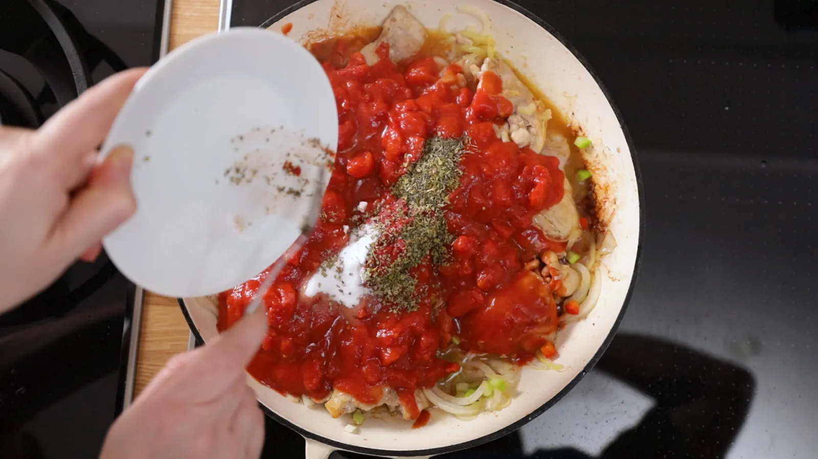 Hands add salt, pepper, and dried herbs from a white plate into a white enameled cast iron pan containing chicken pieces, diced tomatoes, and onions.