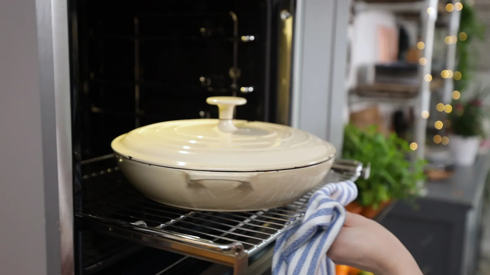 A hand, holding a blue and white striped oven mitt, slides a white enameled cast iron pot with a lid into a hot oven.