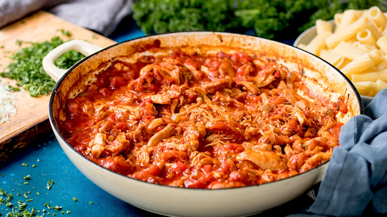 A white enameled cast iron pan filled with shredded chicken in a bubbling red tomato sauce, with cooked pasta and a blue napkin nearby.