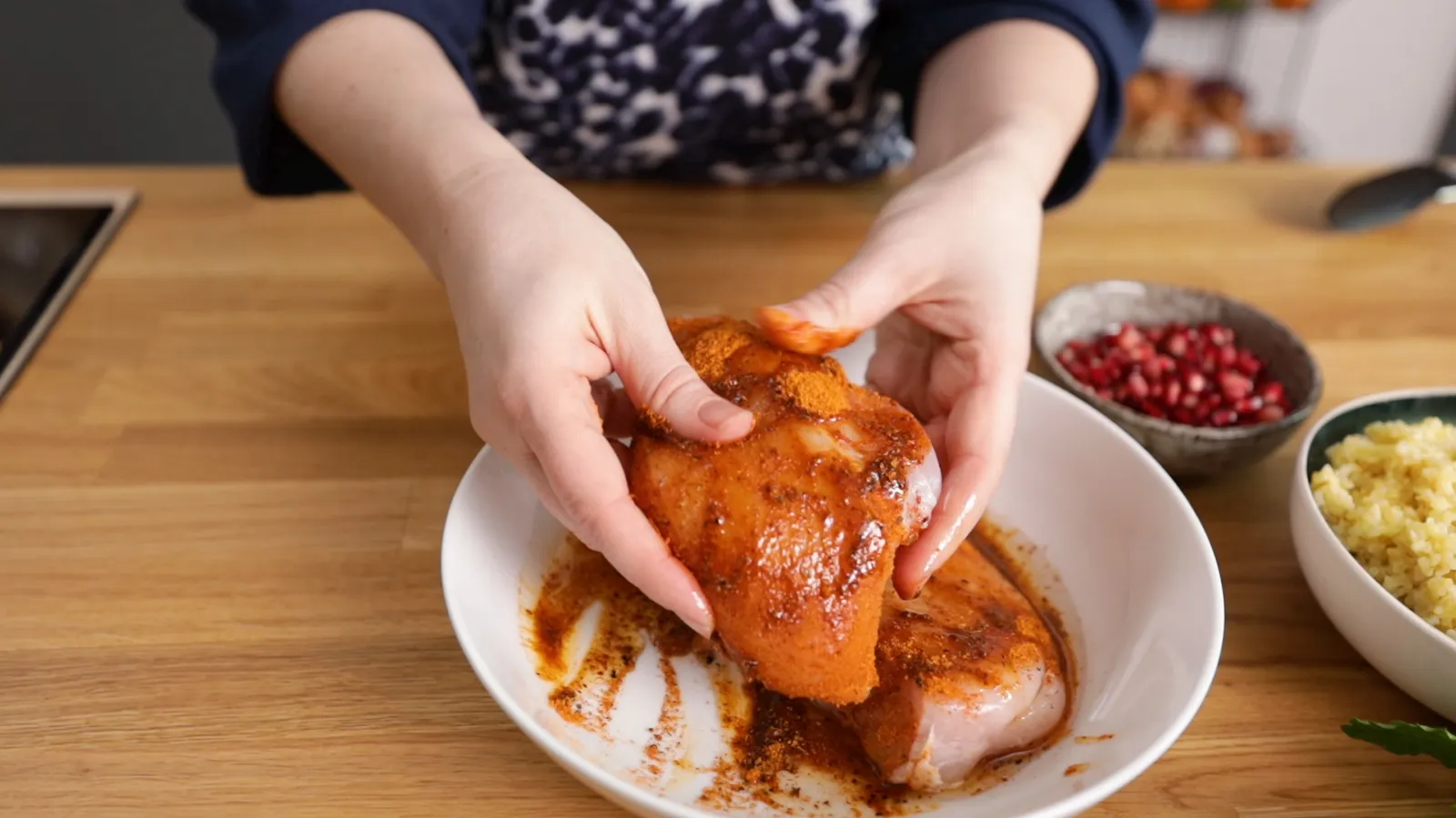 Hands coat raw chicken breasts with a red spice marinade in a white bowl.