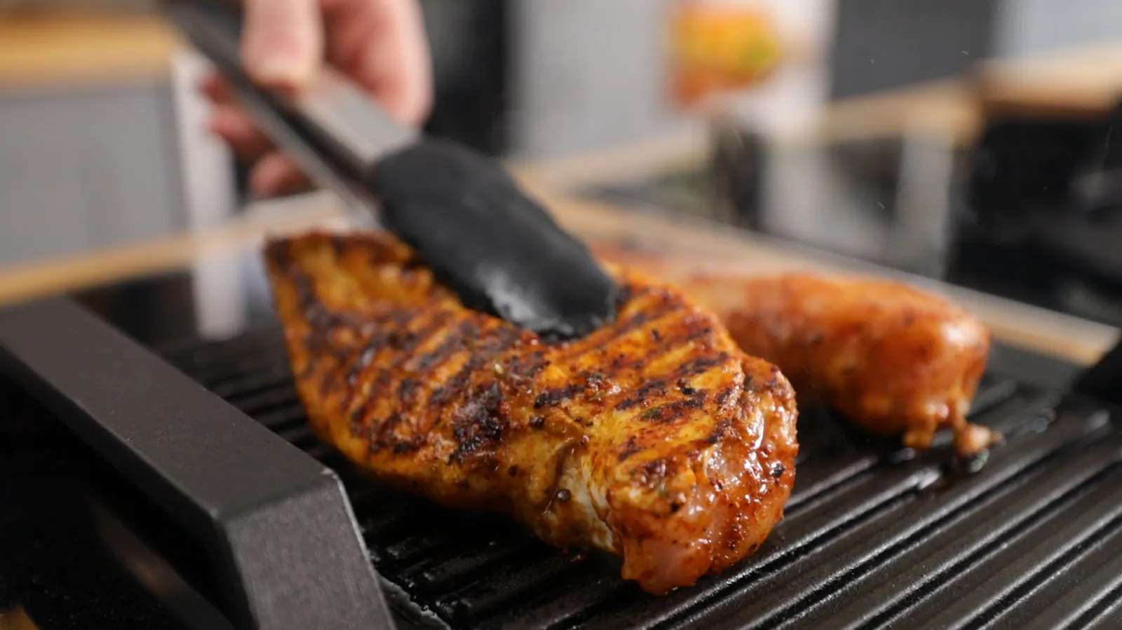 A hand uses tongs to flip a grilled chicken breast on a hot grill pan, showing prominent grill marks, next to a sausage.