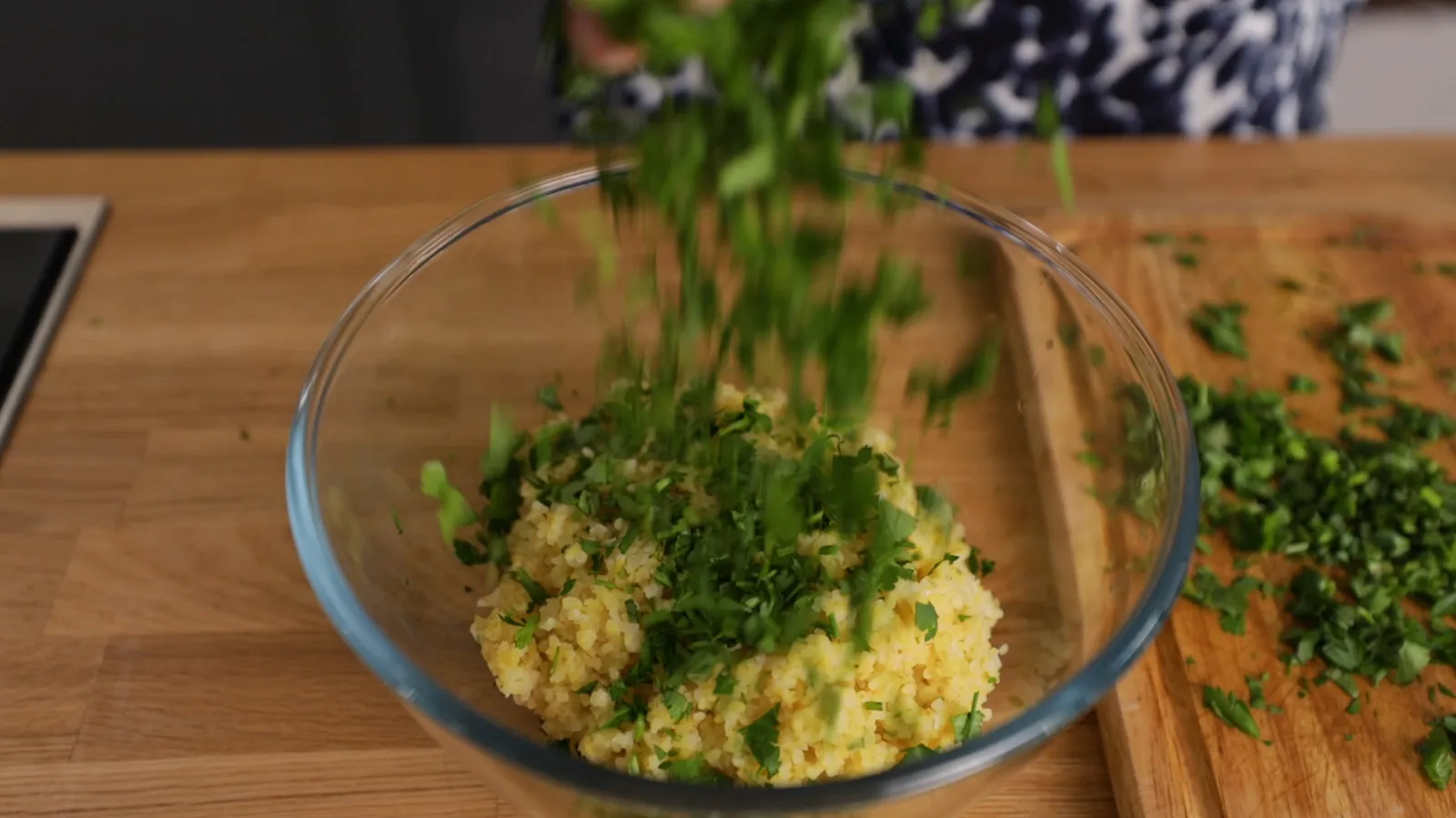 Chopped fresh parsley is sprinkled into a clear glass bowl containing cooked couscous.