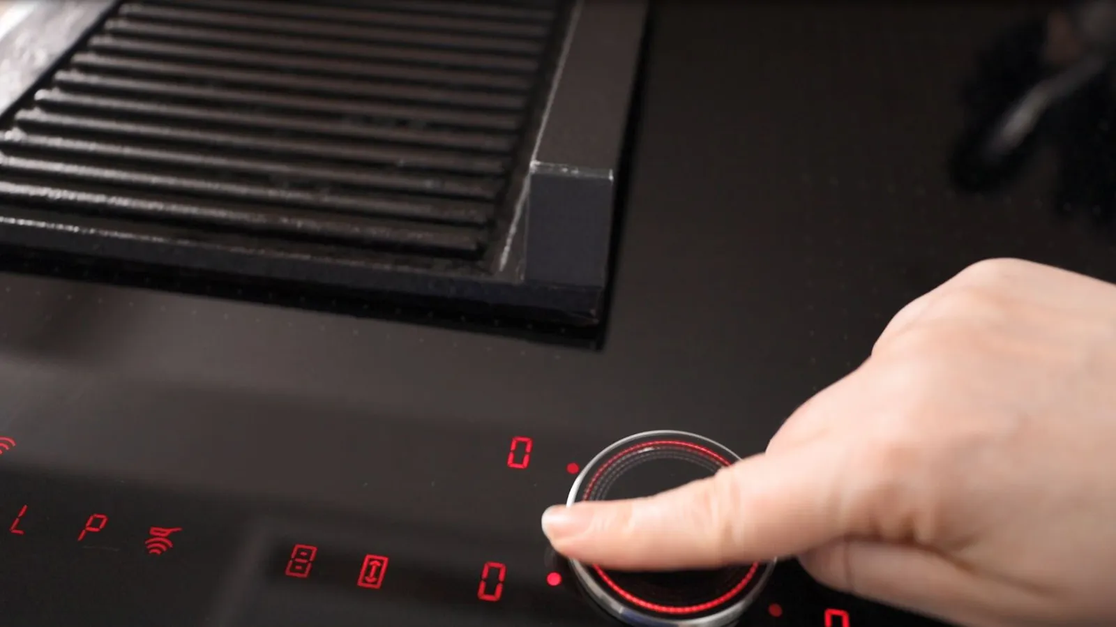 A finger adjusts the illuminated red digital control dial of a black induction cooktop, showing various settings.