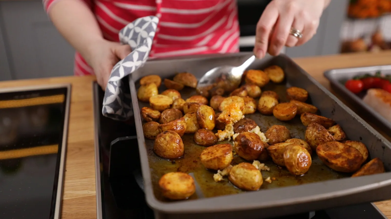A close-up of a baking tray filled with golden-brown roasted potatoes, being stirred with a spoon, with a checkered oven mitt holding the tray.