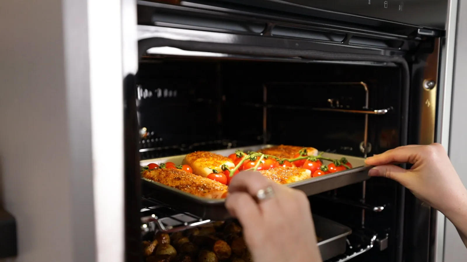 Hands carefully slide a baking tray with seasoned fish fillets and cherry tomatoes into a hot oven, with another tray of roasted potatoes already inside.
