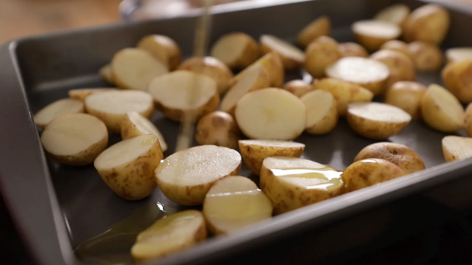 A close-up of halved small potatoes on a baking tray, being drizzled with olive oil.