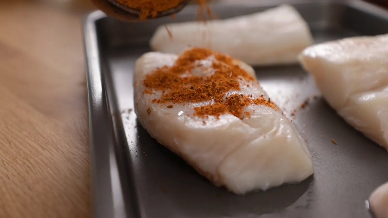 Image 5: A close-up of raw white fish fillets on a baking tray, being generously sprinkled with red seasoning.