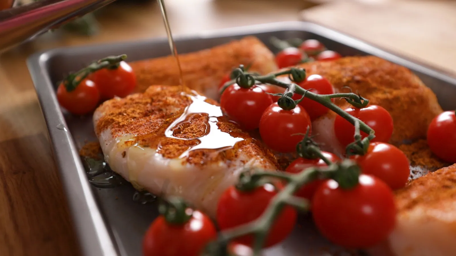 A close-up of raw fish fillets and cherry tomatoes on the vine on a baking tray, being drizzled with olive oil.