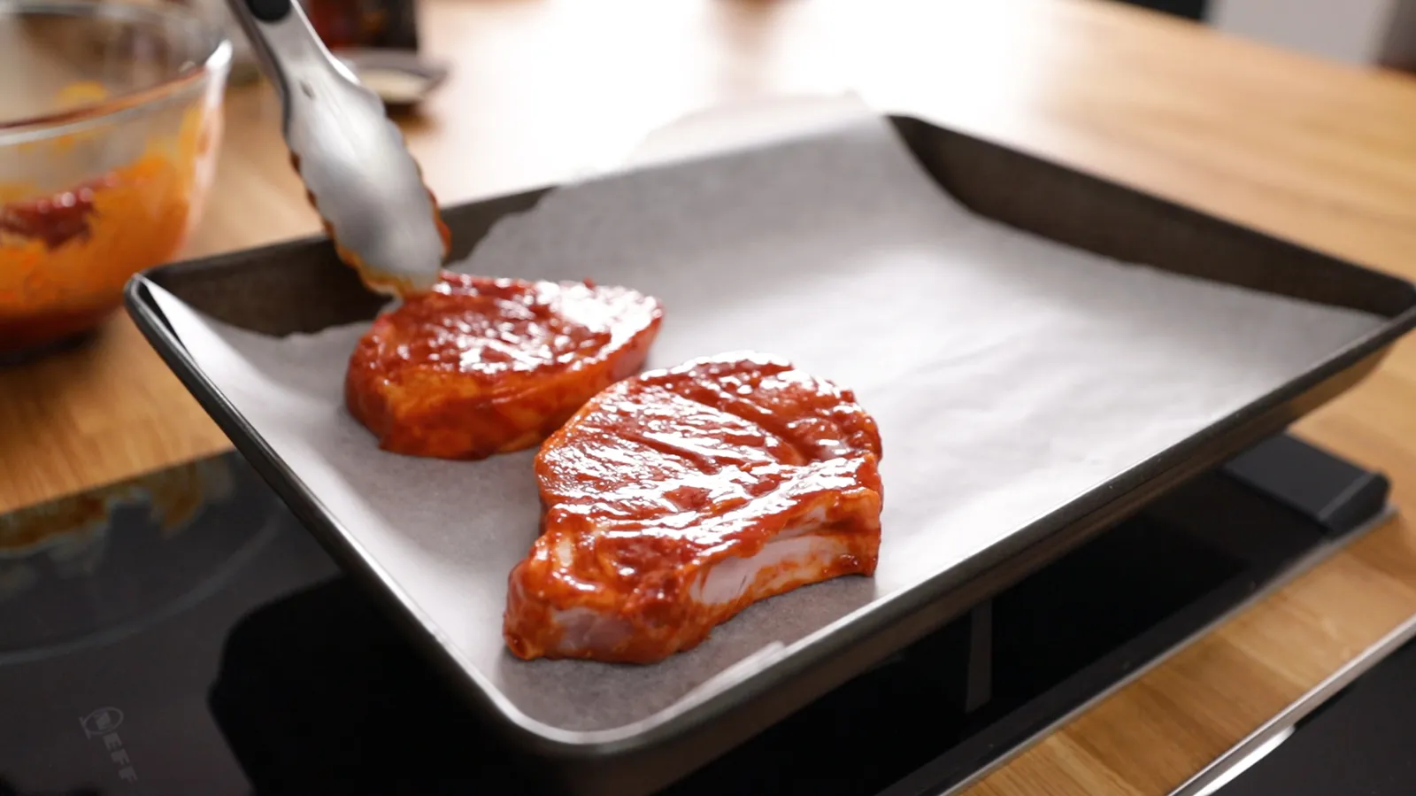 Hands use tongs to place two marinated pork chops onto a baking tray lined with parchment paper.