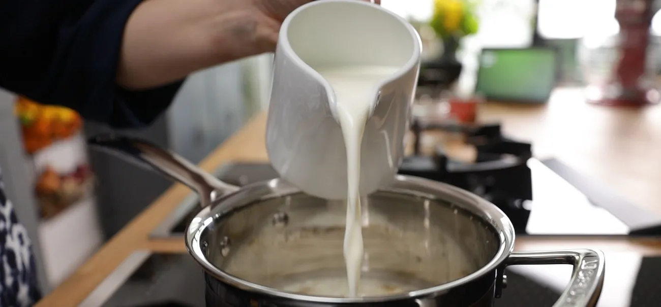 A hand pours white liquid (likely milk or cream) from a white pitcher into a stainless steel saucepan on an induction cooktop.