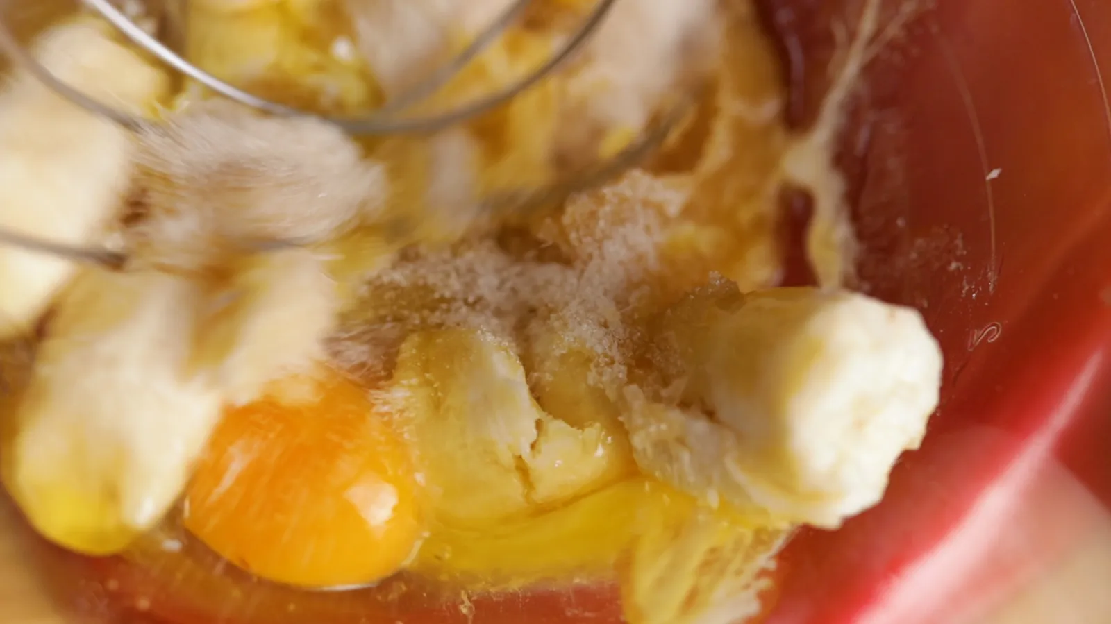 A close-up of a mixing bowl with mashed bananas, a cracked egg, and sugar, being whisked together.