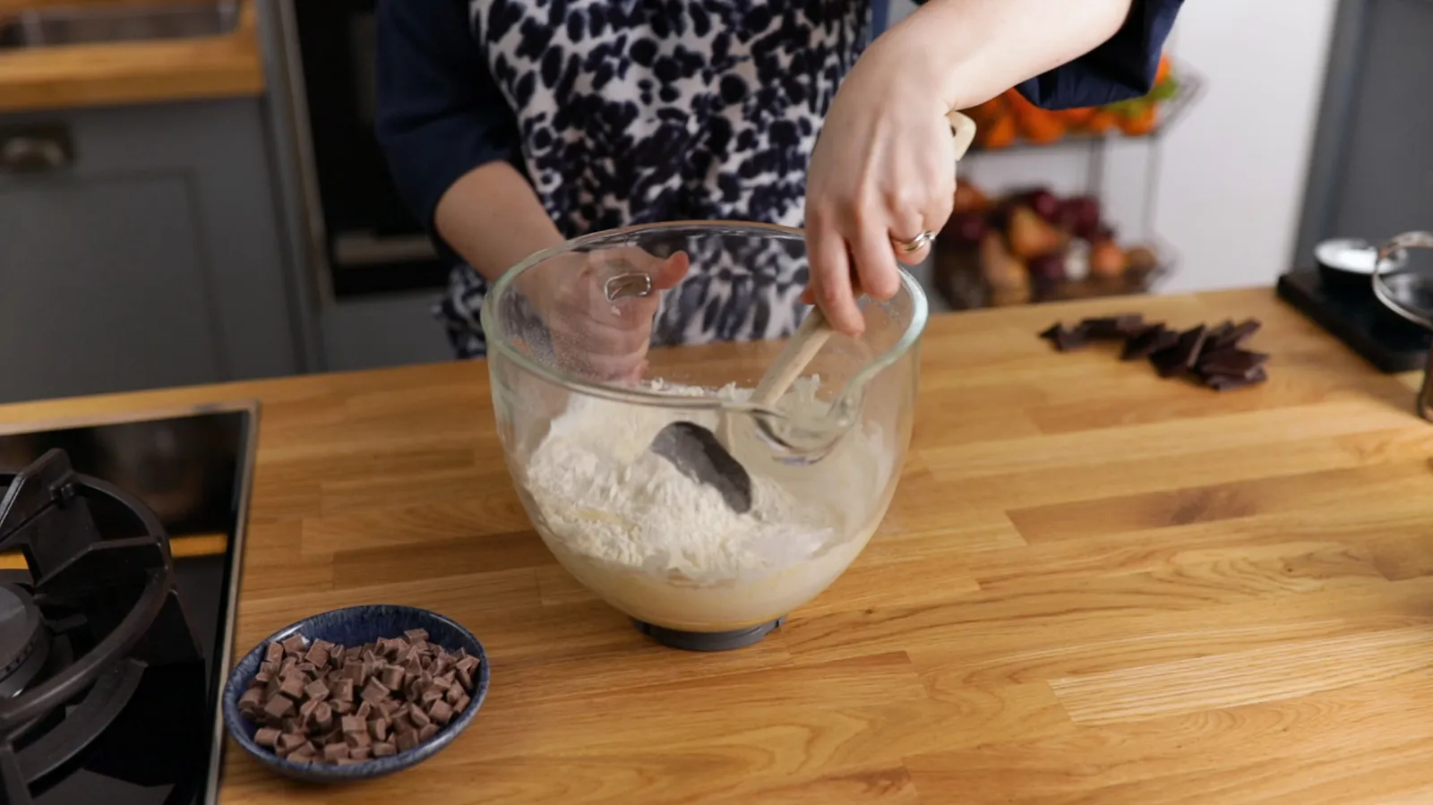 ands use a wooden spoon to mix white flour in a large clear glass bowl on a wooden countertop, with chocolate chips nearby.