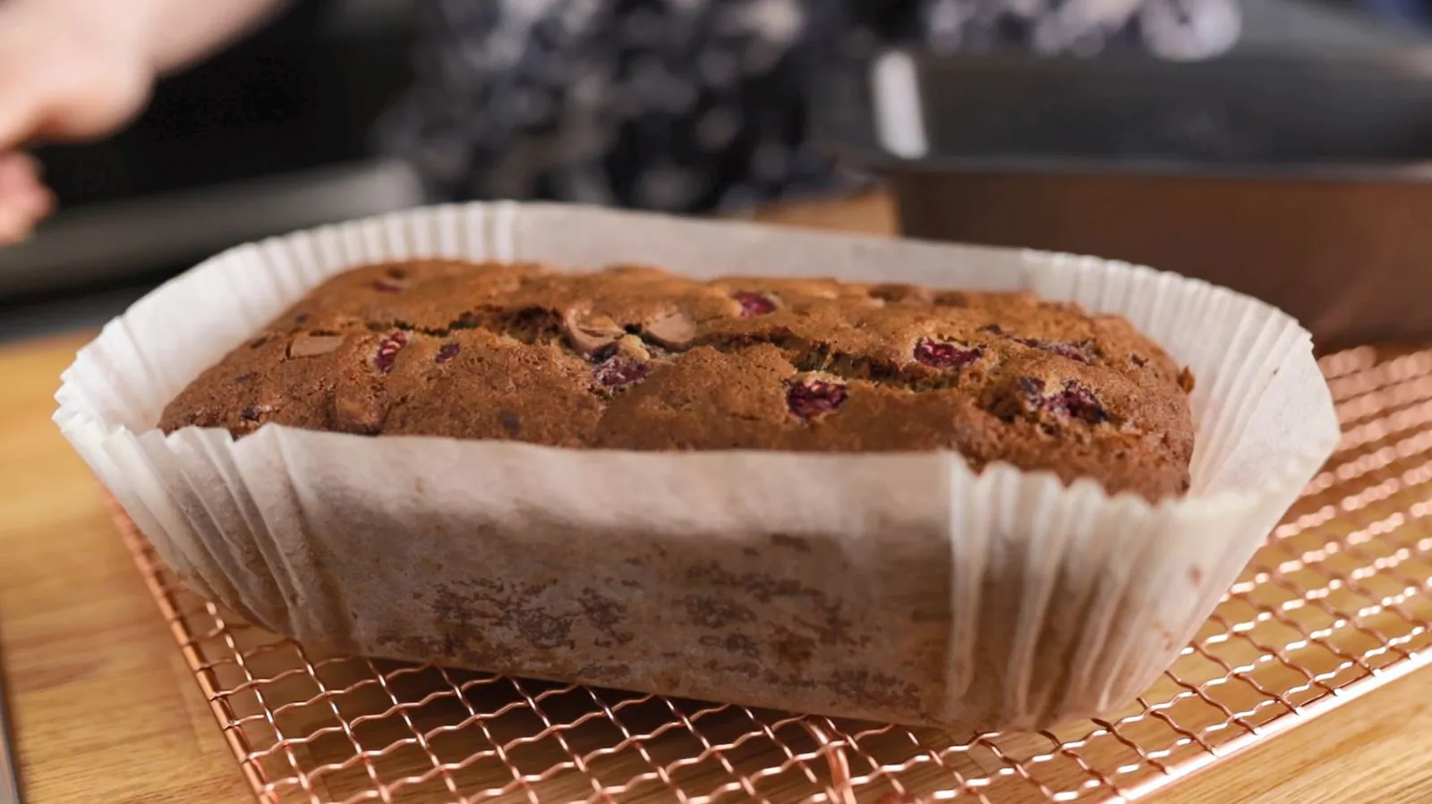 A freshly baked loaf cake, golden brown and studded with red fruit and chocolate chips, rests in its parchment paper liner on a copper wire rack.