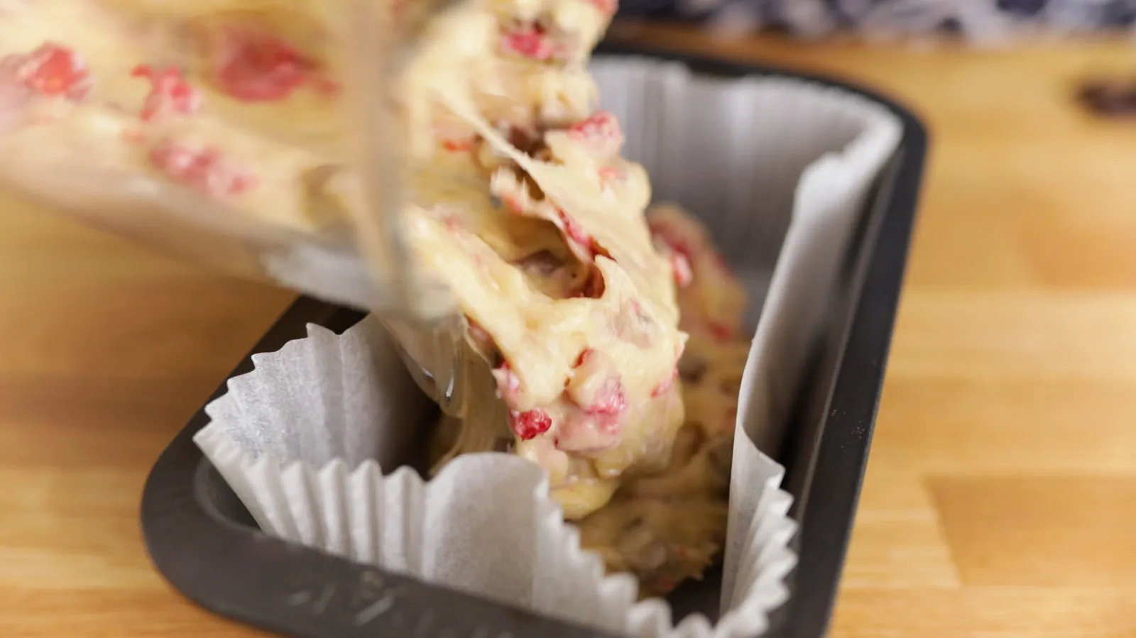 Thick, lumpy batter with red fruit pieces is poured from a clear plastic bag into a loaf pan lined with white parchment paper.