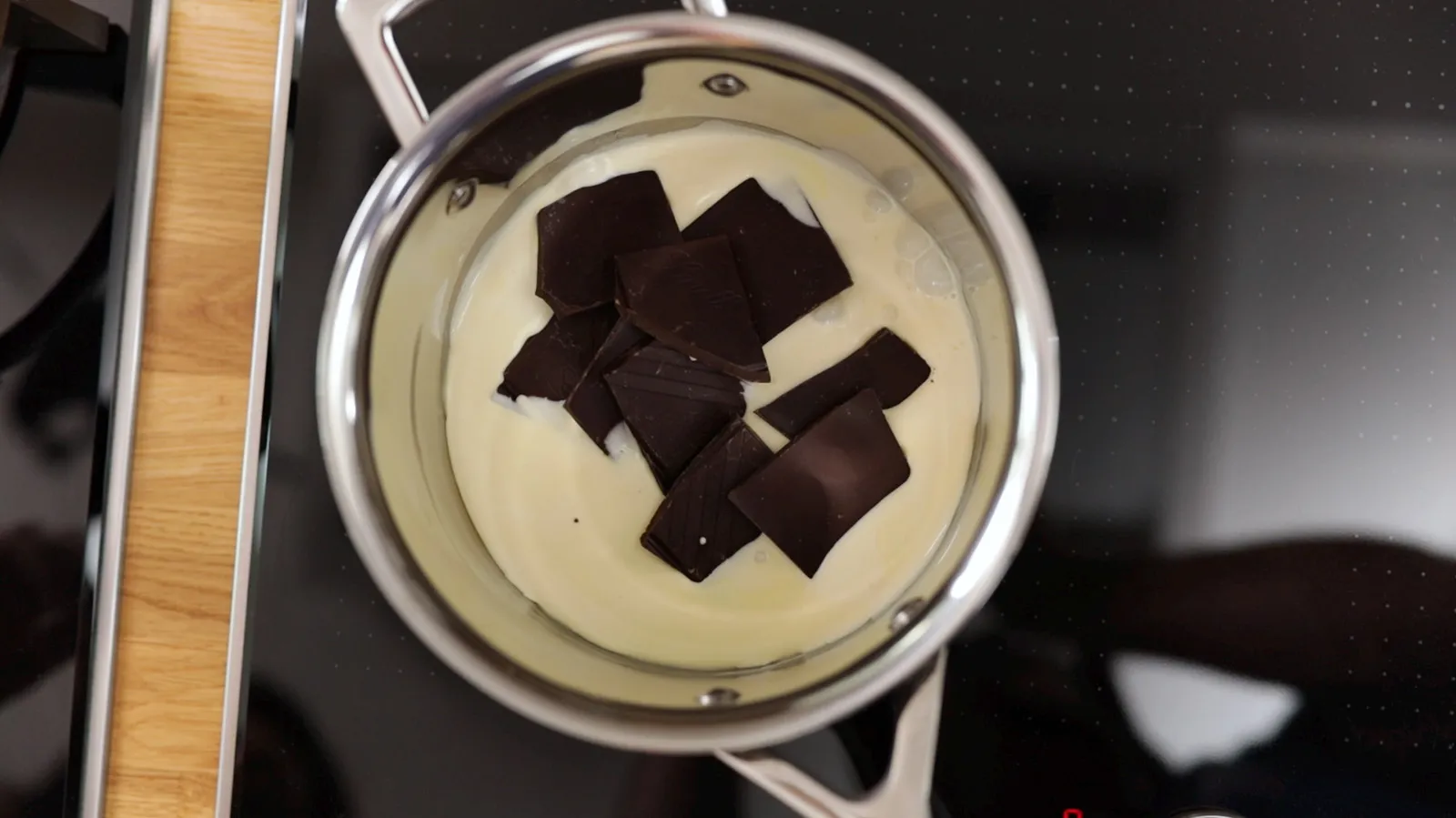 An overhead view of a stainless steel saucepan on an induction cooktop, containing white cream and dark chocolate pieces, melting together.