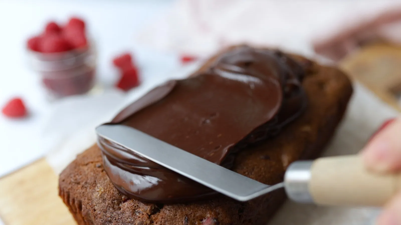 A hand uses a metal spatula to spread thick, melted dark chocolate frosting over a baked loaf cake.