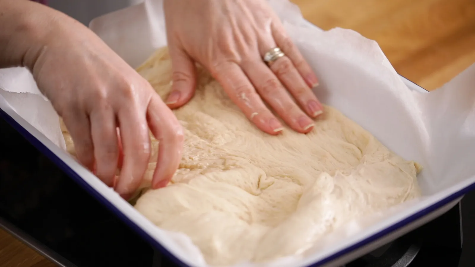 Hands gently press and spread a raw focaccia dough into a white and blue enamel baking dish lined with parchment paper.