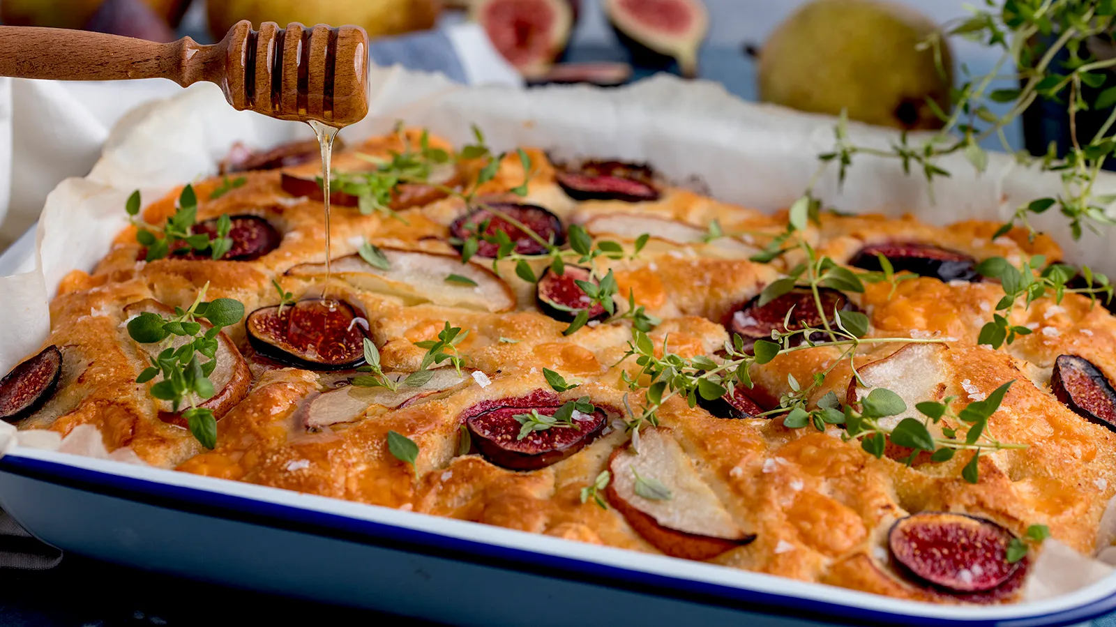 A honey dipper drizzles honey over a golden-brown focaccia bread topped with sliced figs, pears, and fresh thyme, baked in a white and blue enamel dish.