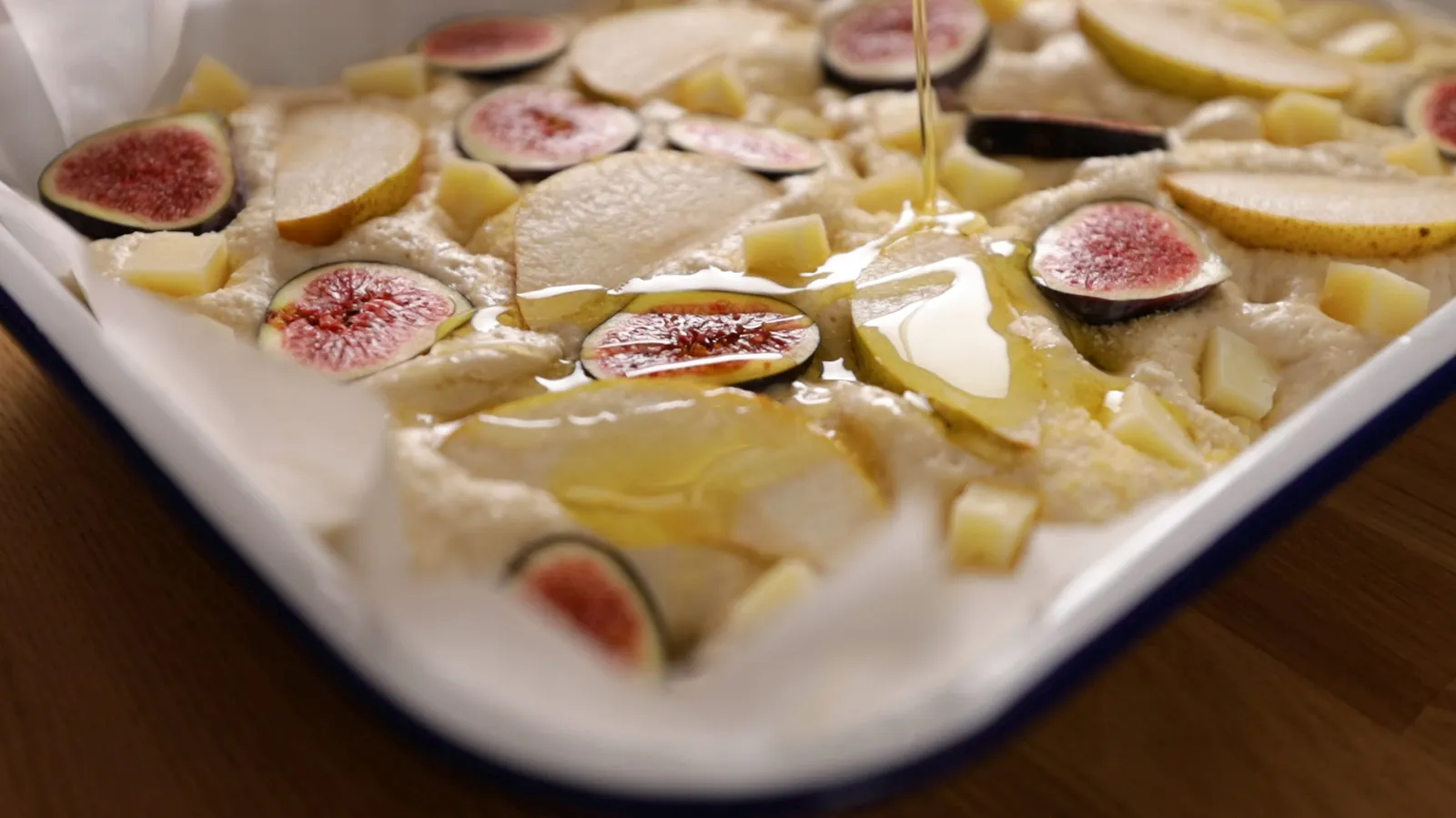 Olive oil is drizzled over a raw focaccia dough topped with sliced figs, pears, and cheese cubes in a white and blue enamel baking dish.
