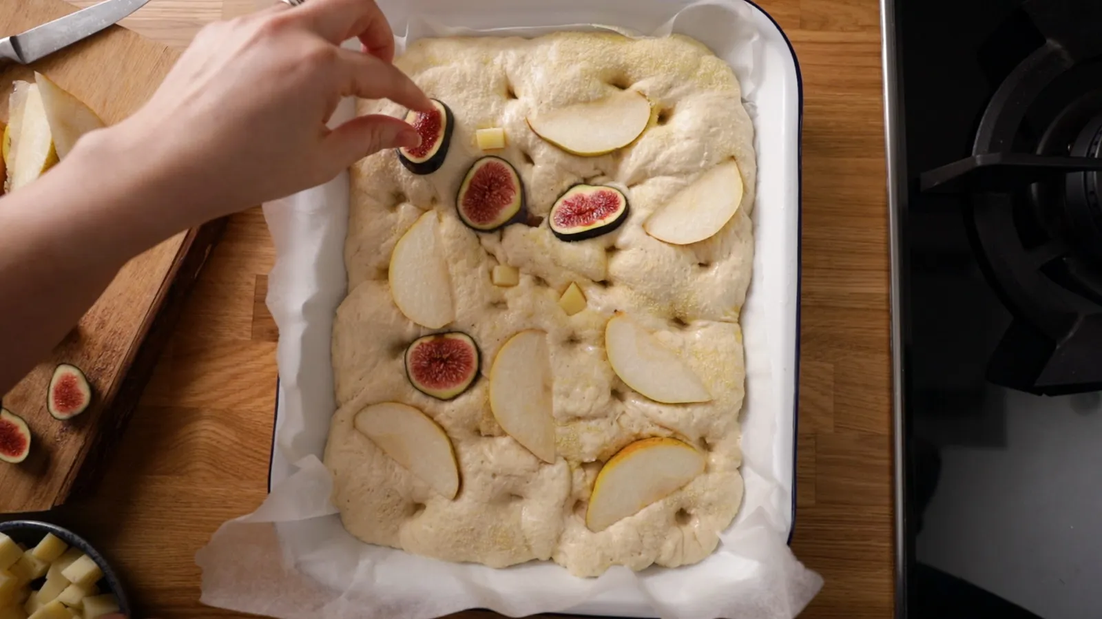 Hands arrange sliced figs and pears on top of a raw focaccia dough in a white and blue enamel baking dish.