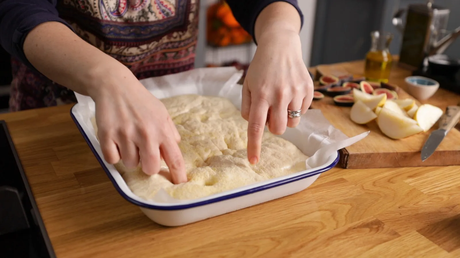 Hands gently press down on a raw focaccia dough in a white and blue enamel baking dish, creating dimples.