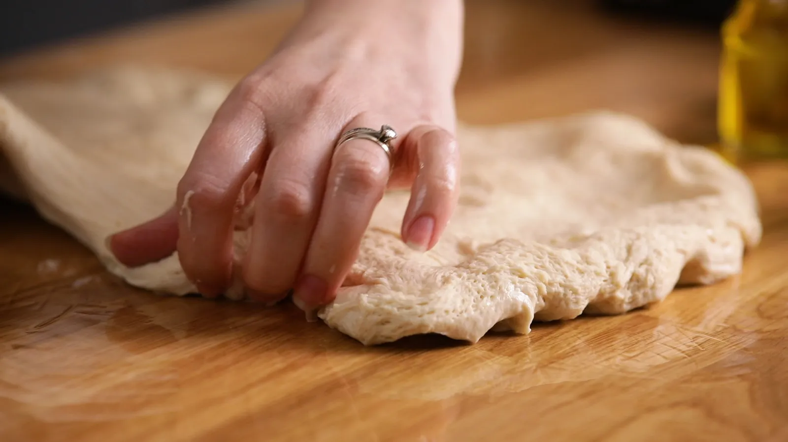 A hand gently stretches and folds a piece of raw focaccia dough on a wooden surface.
