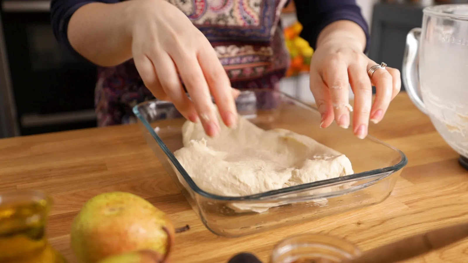 Hands transfer a large piece of raw focaccia dough into a clear glass baking dish.