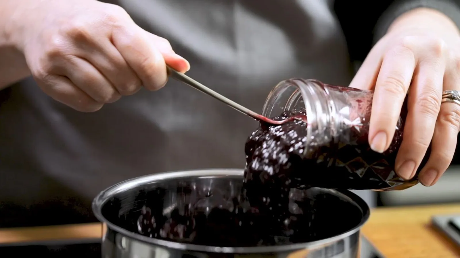 Hands use a spoon to transfer dark cherry jam from a glass jar into a stainless steel saucepan.