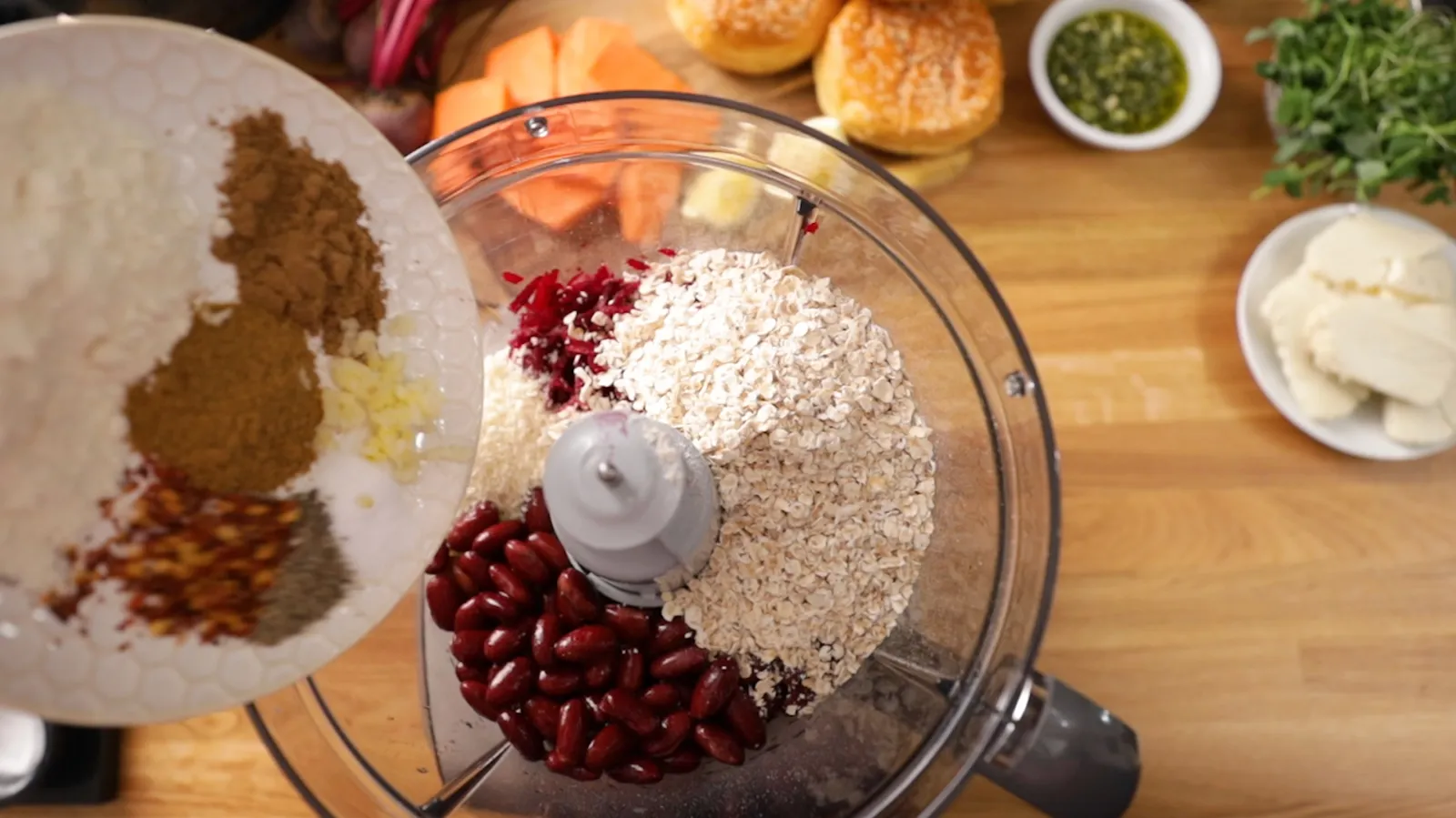 An overhead shot of a food processor bowl filled with red beans, oats, and chopped beets, as a hand adds spices from a white plate.