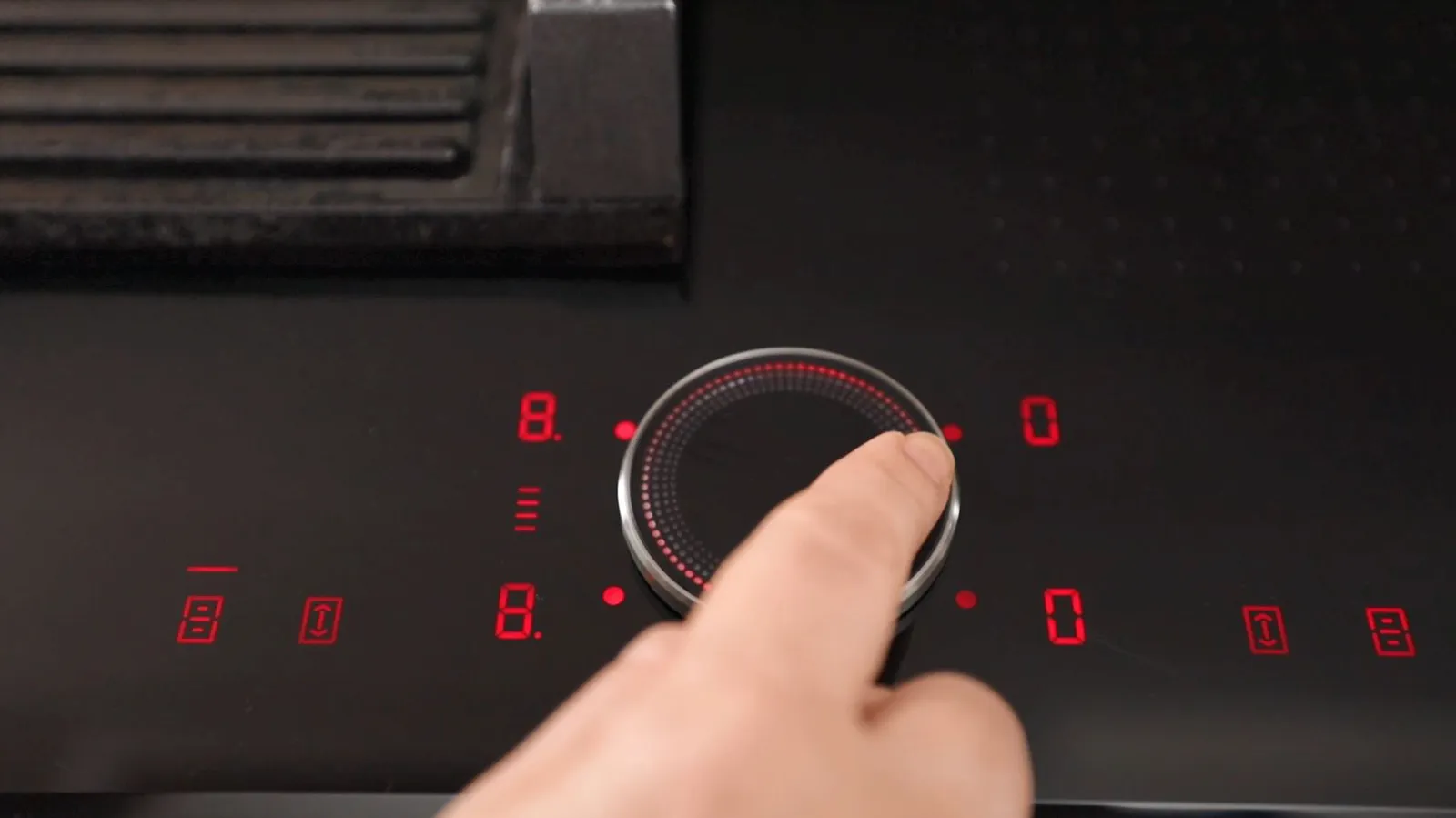 A finger adjusts the illuminated digital control dial of a black induction cooktop, showing red numbers and settings.