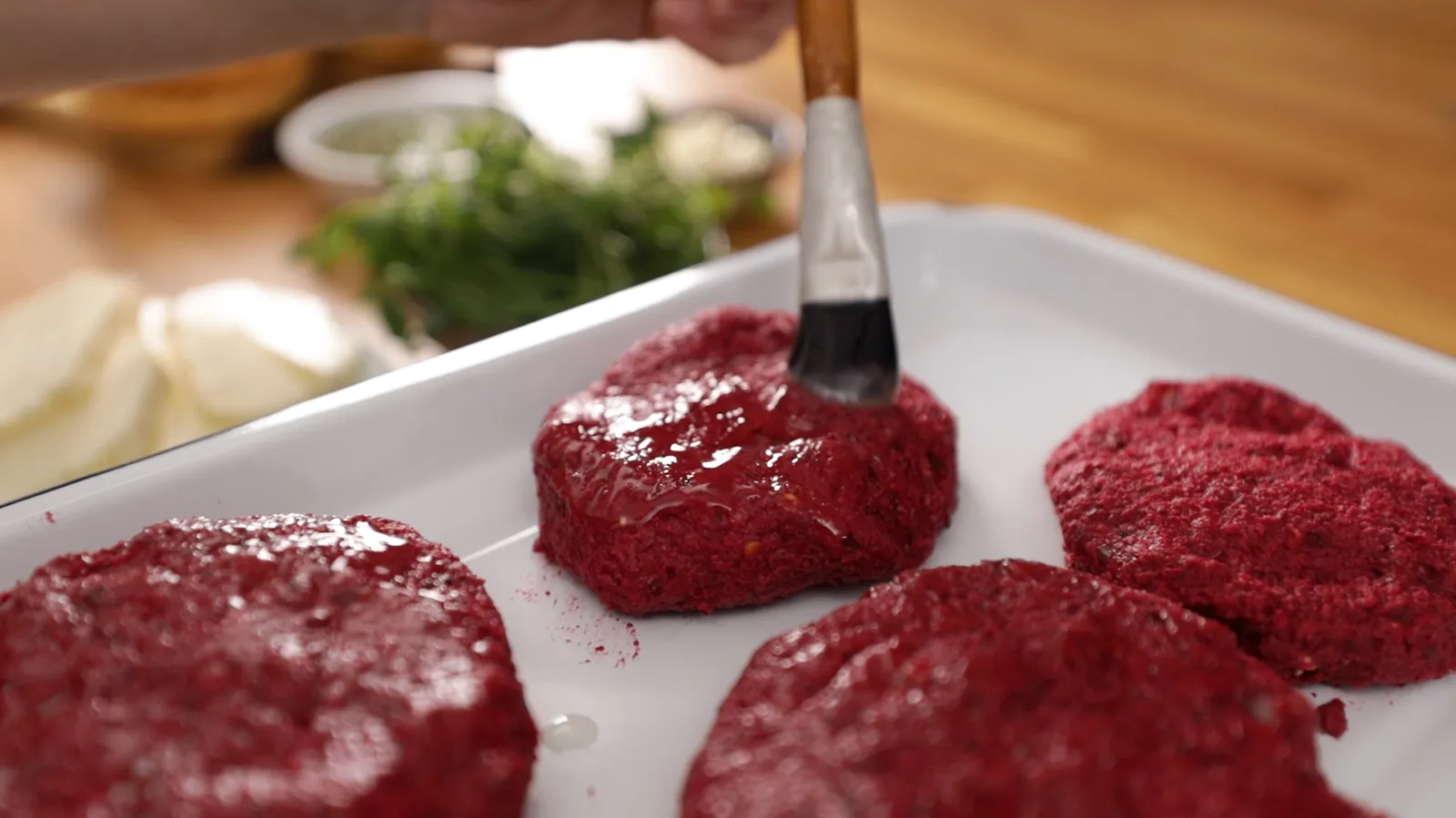 A hand brushes oil onto raw, round beet patties arranged on a white baking tray.