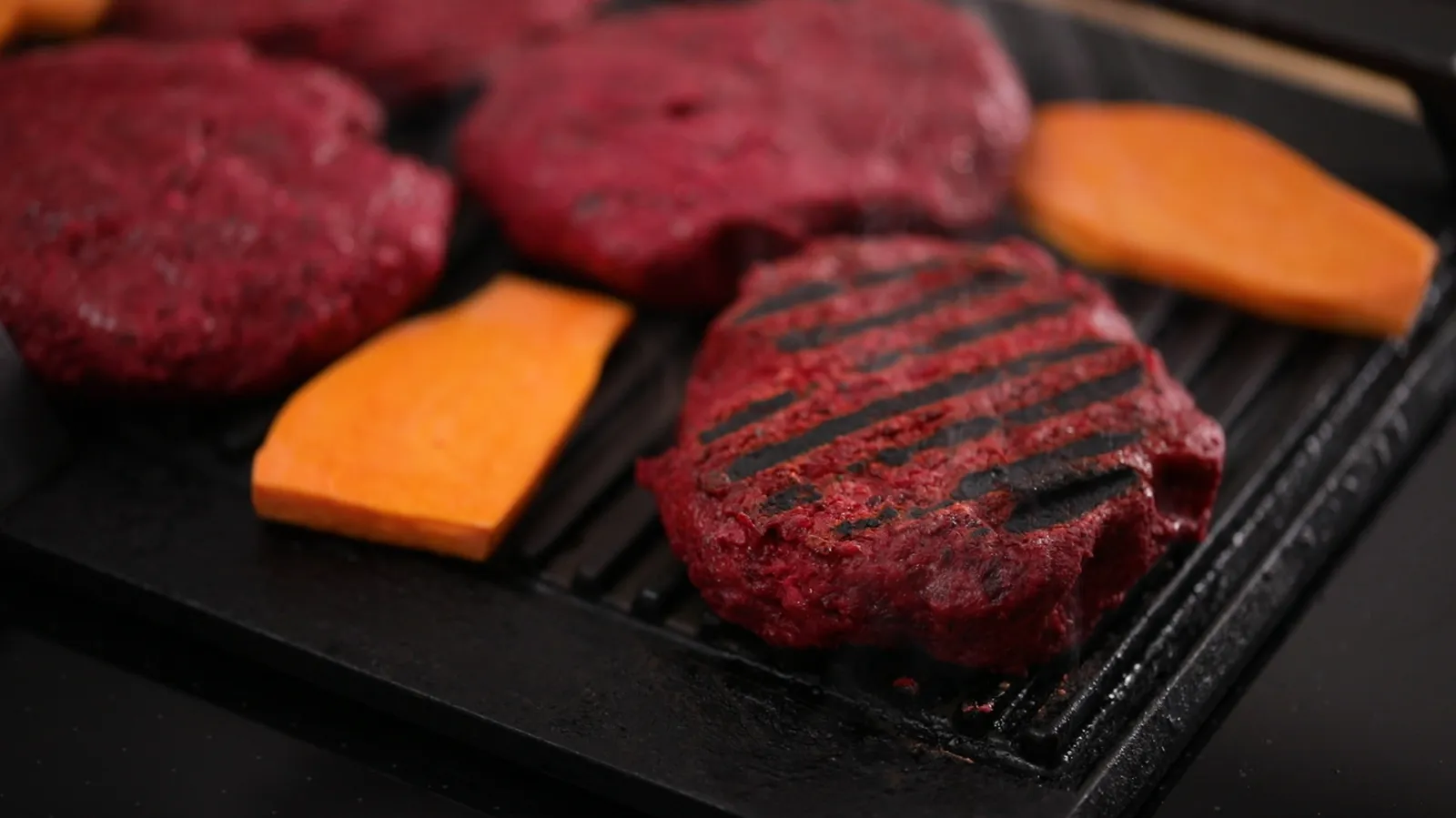 A close-up of a hot grill pan with a cooked beet patty showing grill marks, alongside raw beet patties and sweet potato slices.