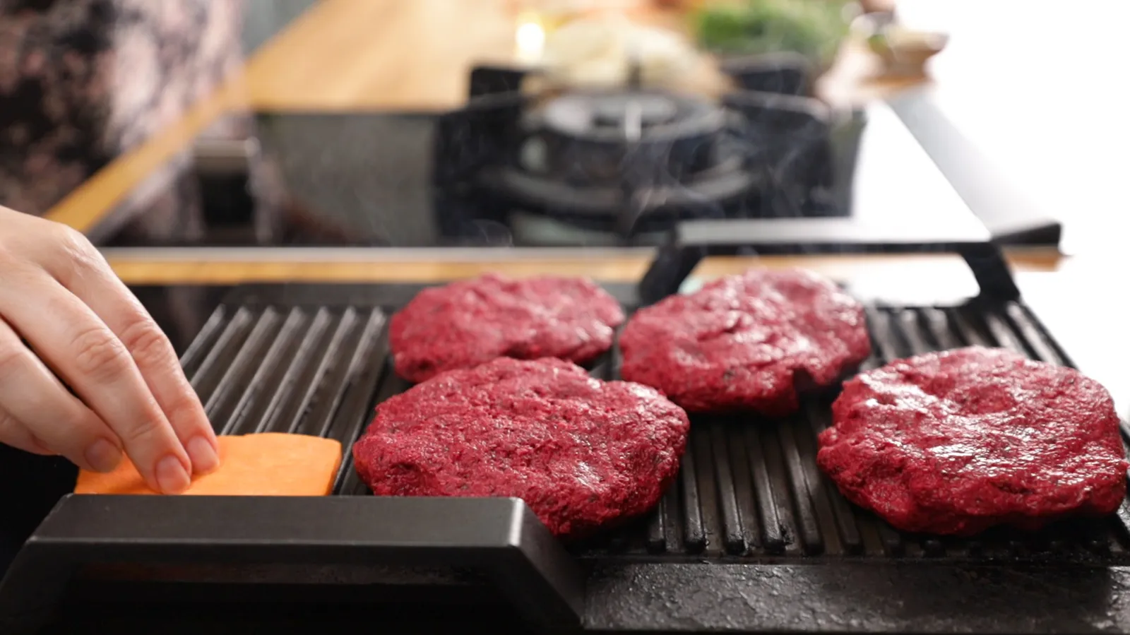 A hand places a raw beet patty onto a hot grill pan on an induction cooktop, next to other patties and sweet potato slices.