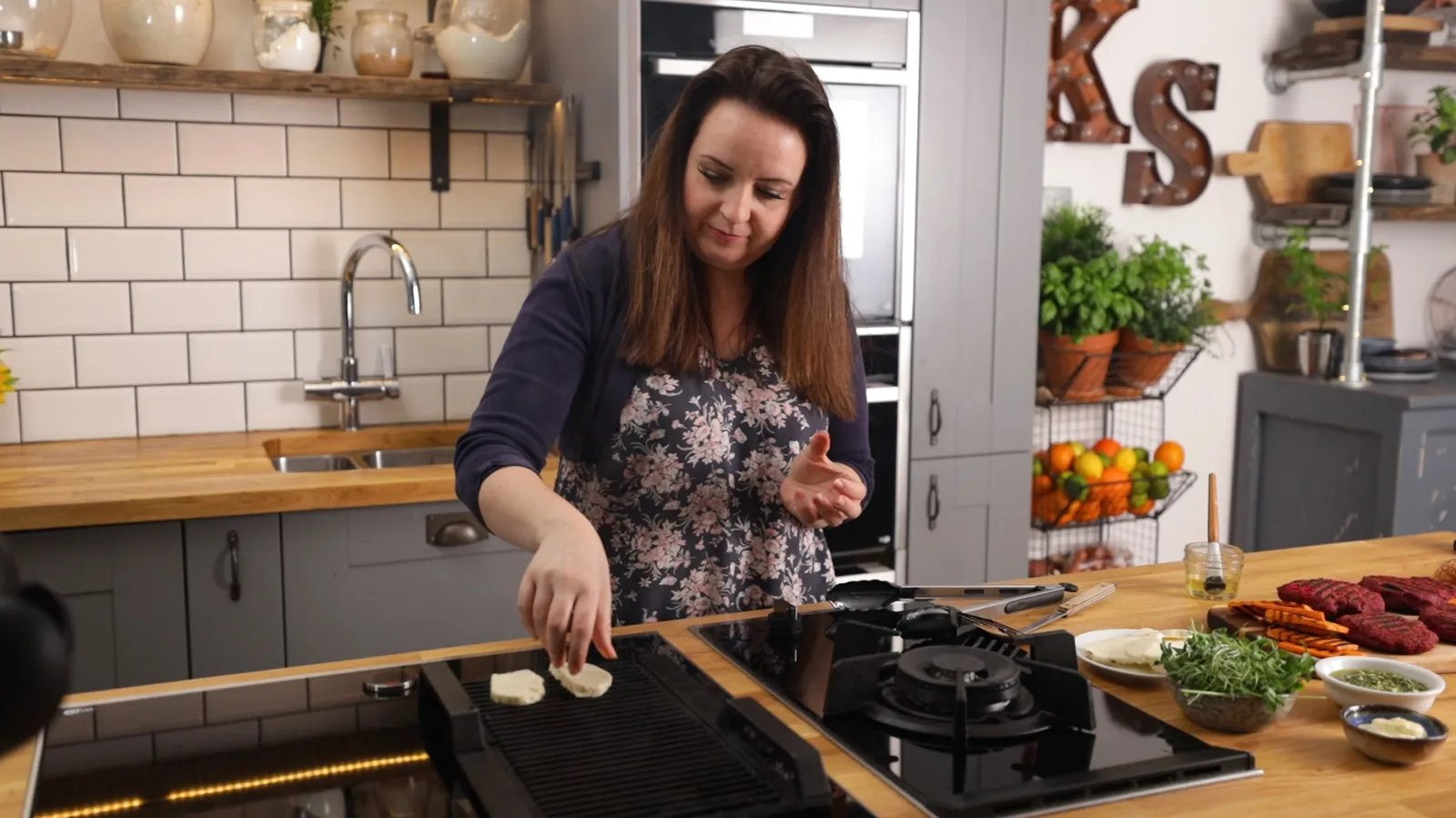 A woman places small pieces of halloumi cheese onto a hot grill pan on an induction cooktop in a modern kitchen.