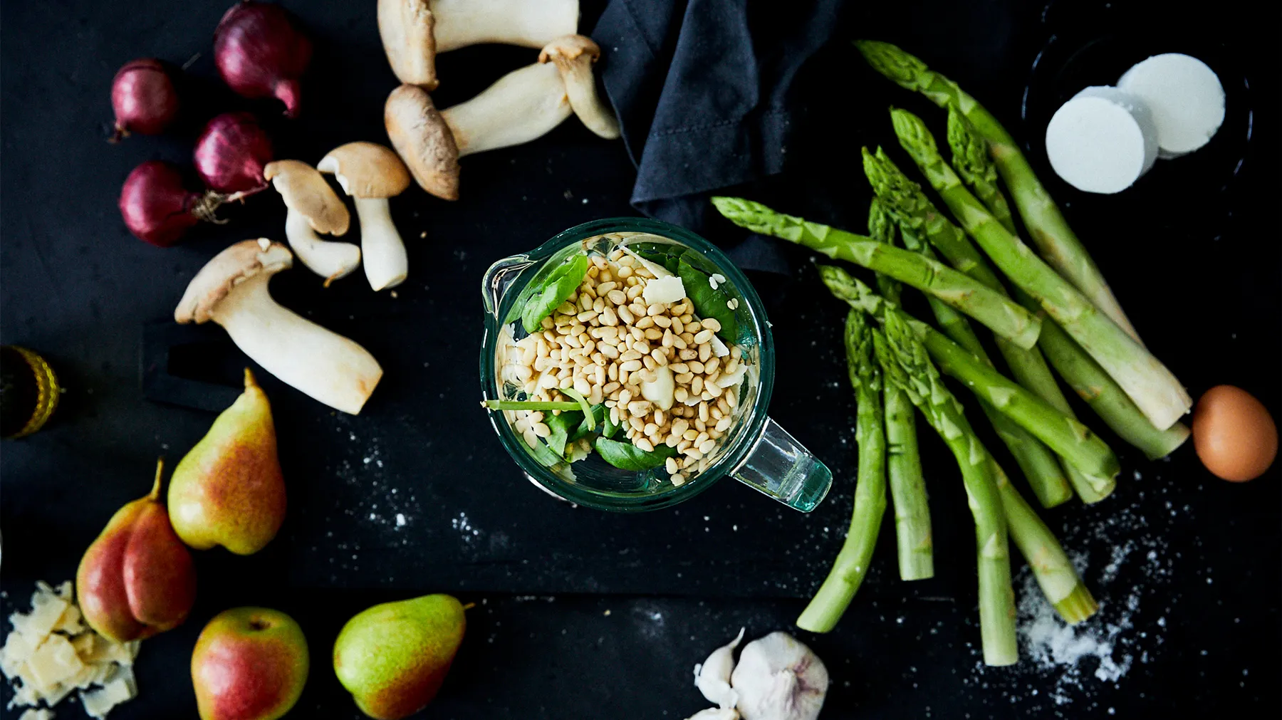 An overhead flat lay of pesto ingredients: a blender filled with fresh basil and pine nuts, fresh asparagus, red onions, mushrooms, pears, cheese, and garlic, all on a dark surface.