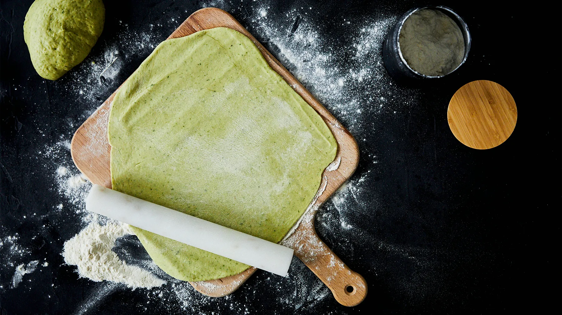 An overhead view of a light green quiche dough rolled out on a wooden cutting board, with a white rolling pin, flour, and a container of green powder on a black surface.