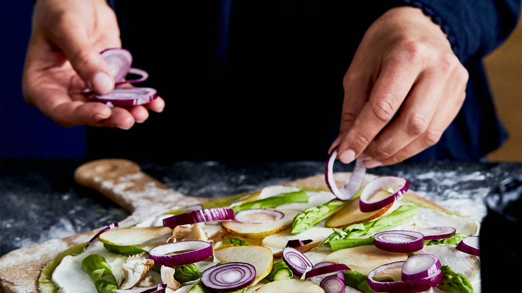 Hands arrange thin slices of red onion on a green pizza dough topped with pears, asparagus, and cheese, on a wooden cutting board.
