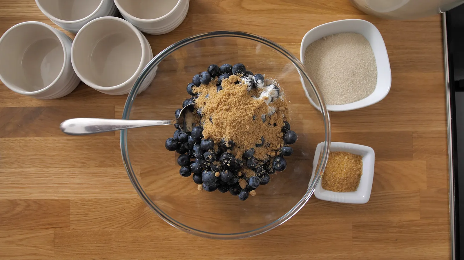 An overhead shot of a clear glass bowl filled with fresh blueberries, brown sugar, and a spoon, surrounded by small white bowls and empty ramekins on a wooden countertop.